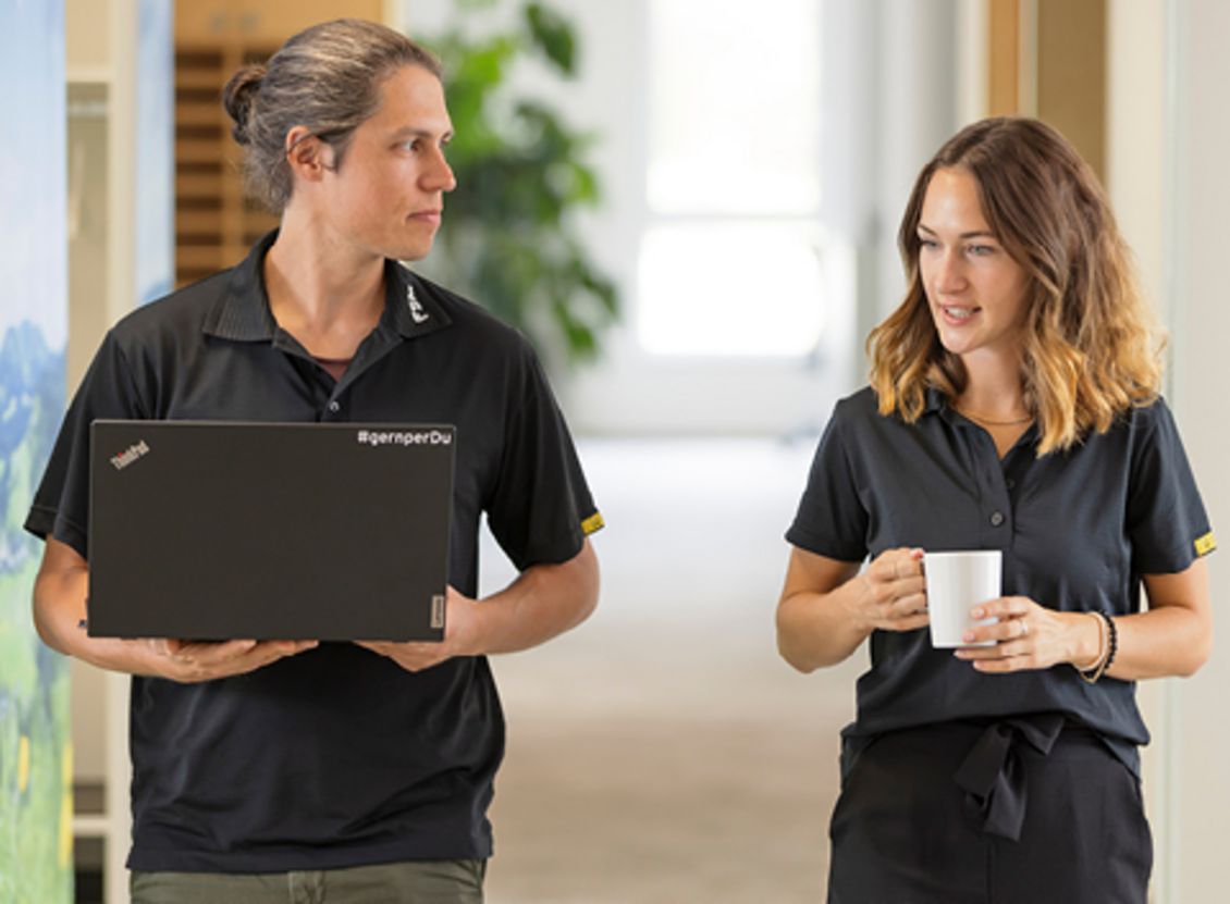 Zwei Mitarbeiter der FSM AG in einem Flur. Der Mitarbeiter links trägt einen Laptop und blickt zur Kollegin, die rechts mit einer Tasse in der Hand steht. Die helle und moderne Umgebung deutet auf eine freundliche und offene Arbeitsatmosphäre hin.